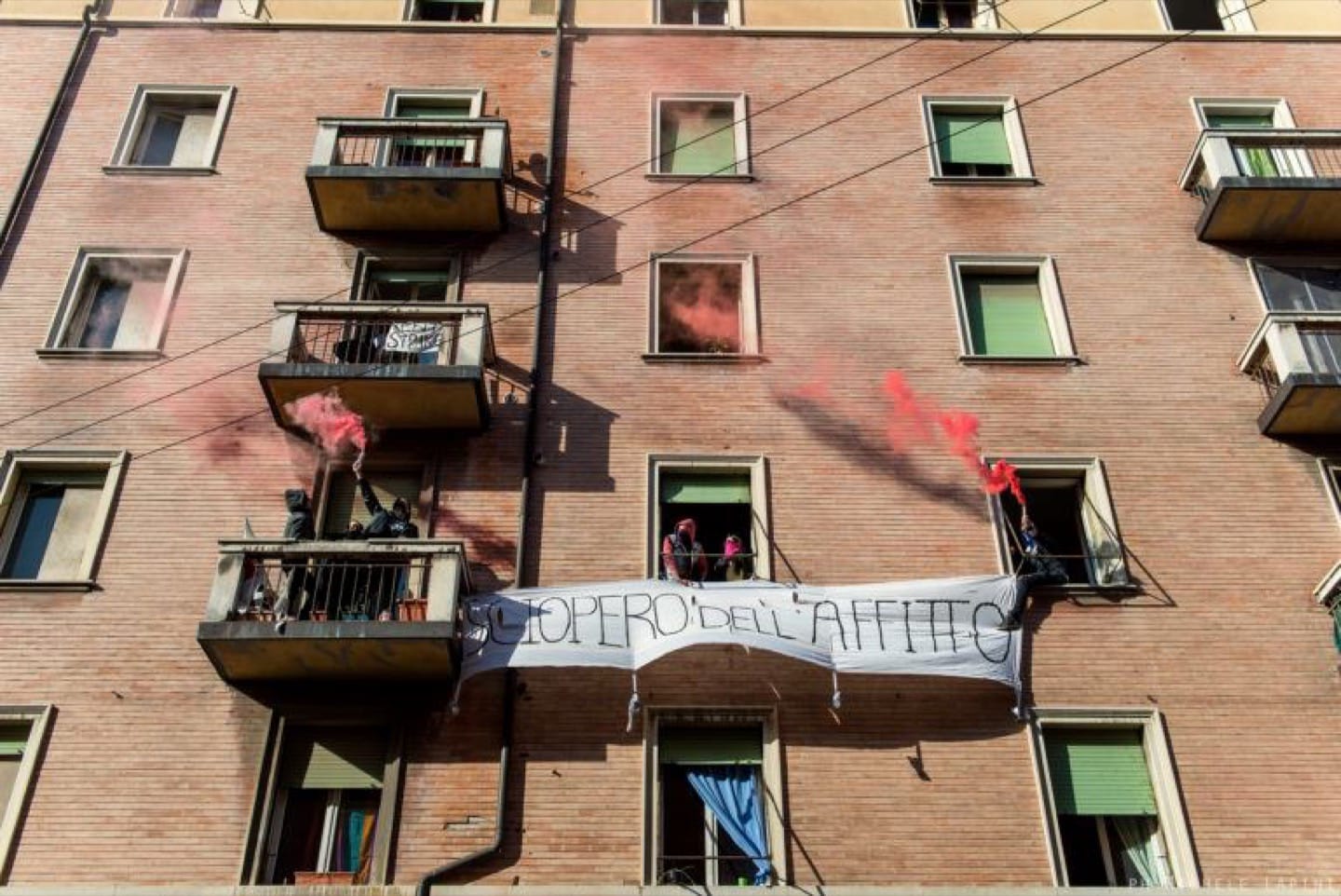 fumogeni rossi si sollevano dalle finestre di una palazzina, uno striscione legge Sciopero dell’affitto